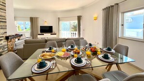 Ein modernes Wohnzimmer mit einem Esstisch aus Glas, gedeckt für das Frühstück, einer Couch und einem Fernseher im Hintergrund.