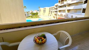 Un balcon avec une table et des chaises, un panier de fruits, et une vue sur une piscine et des bâtiments.