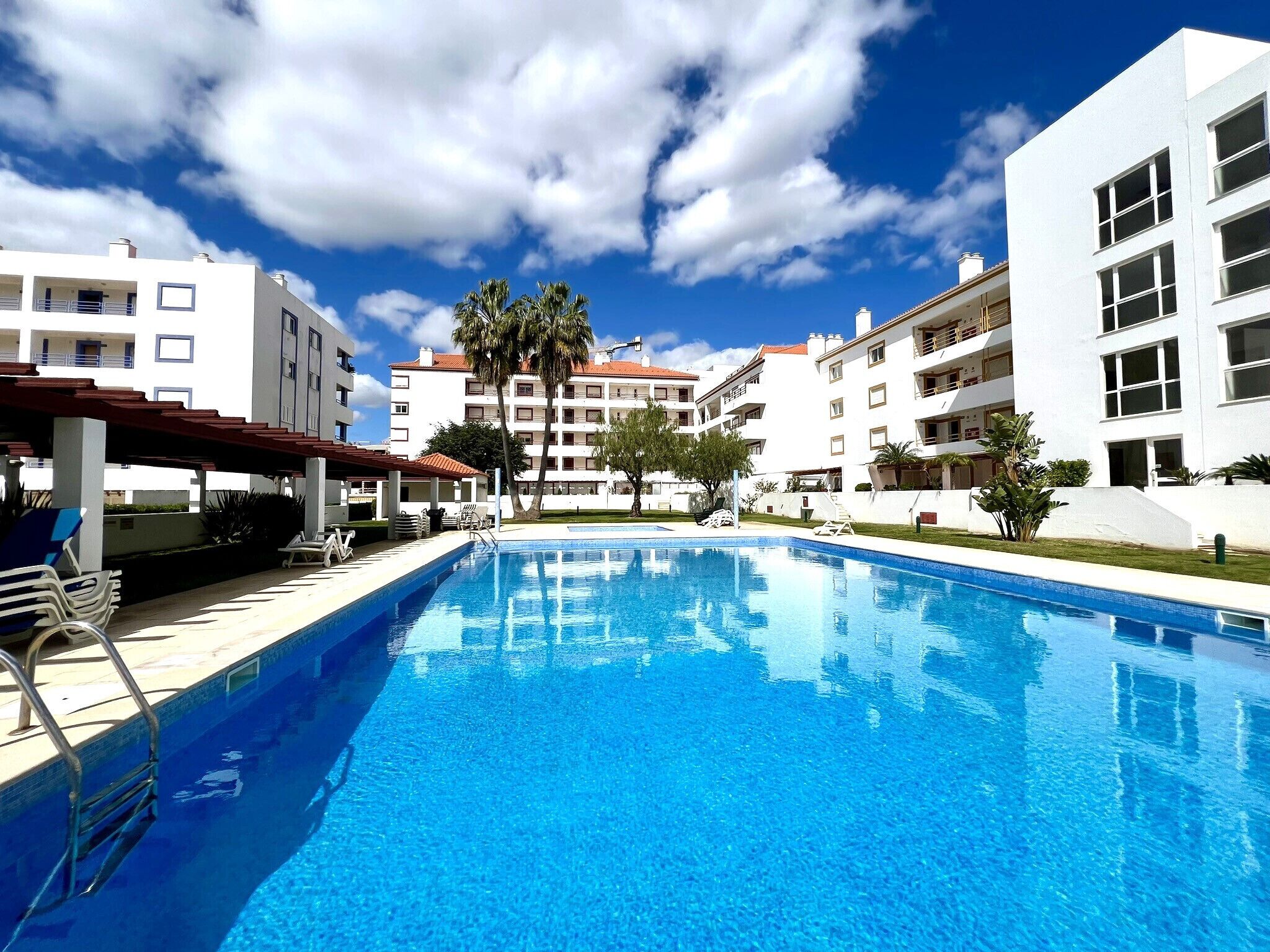 A swimming pool with lounge chairs and a covered area, surrounded by white buildings with balconies.