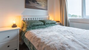 Een slaapkamer met een bed, nachtkastje, lamp, raam met gordijnen en een schilderij aan de muur.