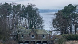 Een rustiek stenen gebouw met een brug over een rivier, omgeven door bomen en een waterpartij op de achtergrond.