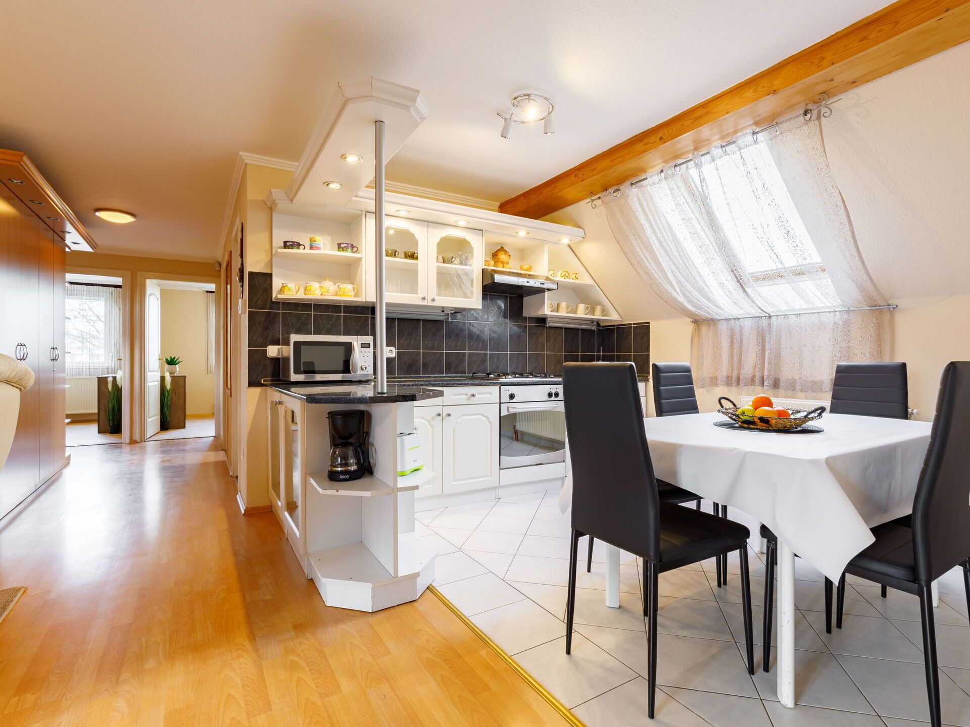 A modern kitchen with white cabinets, a microwave, and a coffee machine. There is a dining area with a table and chairs.