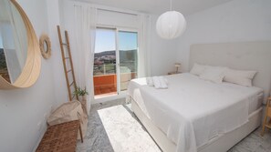 Ein Schlafzimmer mit einem großen Bett, einem Nachttisch, einer Lampe, einem Spiegel und Blick ins Freie.