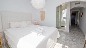 Ein helles Schlafzimmer mit einem Bett, einer Nachttischlampe, einem Wandbehang aus Makramee und Blick auf einen Balkon durch eine geöffnete Tür.