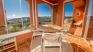 Ein Balkon mit rundem Tisch und Korbstühlen, der Blick auf eine hügelige Landschaft gewährt.