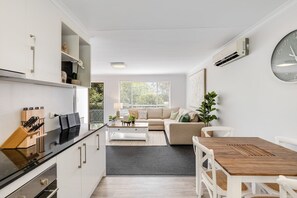 A modern kitchen with white cabinets, a black countertop, and a built-in oven. There is a dining area with a wooden table and white chairs. A large window with blinds is visible in the background.