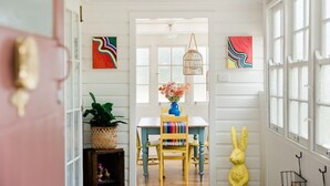 Un pasillo con una puerta blanca, una mesa con un jarrón de flores, una escultura de conejo amarillo y una lámpara colgante.