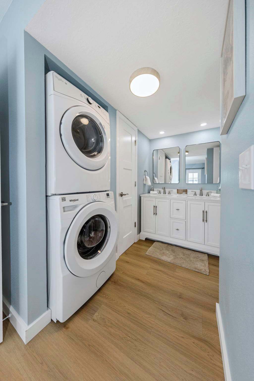 Pequeña lavandería con lavadora y secadora de carga frontal, armarios blancos y un espejo sobre el lavabo.