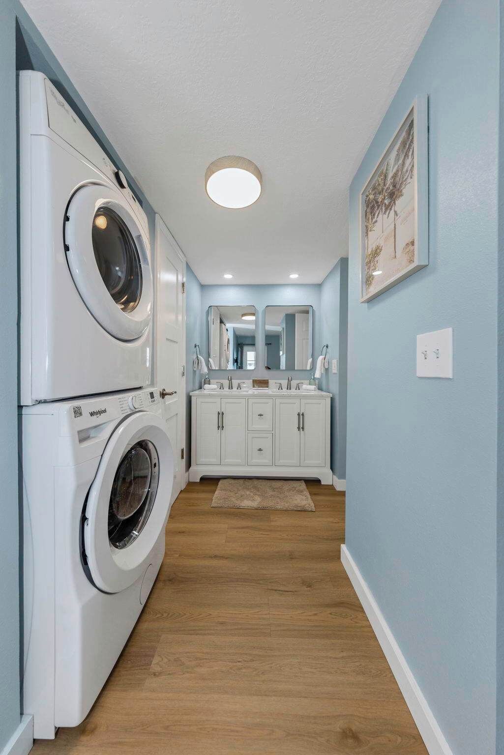 A laundry area with stacked washers and dryers, a white vanity with a mirror, and a wooden floor.
