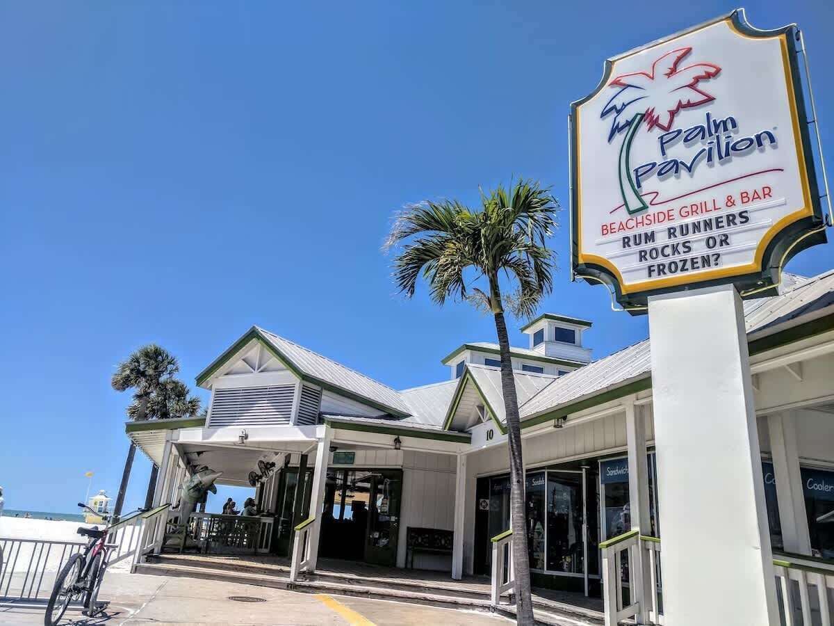 Un restaurante frente a la playa, con una palmera, un letrero que dice 'Palm Pavilion' y vista al mar.