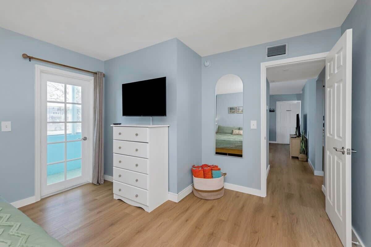 Una habitación con puerta corredera de vidrio, un aparador blanco, un televisor de pantalla plana, un espejo y una cesta con ropa doblada.