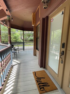 Un porche abrité avec une balustrade en bois, une porte sur laquelle est posé un paillasson avec le mot « bonjour », et une vue sur une cour arrière avec des arbres et une clôture.