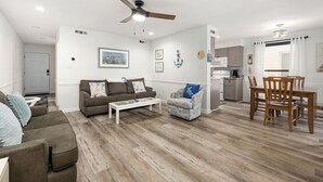 A living room with a sofa, armchair, coffee table, and dining table with chairs. There is a ceiling fan, a kitchen area with cabinets and a microwave, and a door leading to another room. - Canalfront 2 Bedroom-Sleeps 6-Laundry-WiFi (Ocean City)