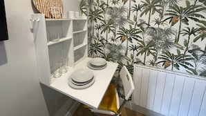 Canto de um quarto com uma estante branca com pratos e copos, um radiador e uma parede com papel de parede de plantas tropicais.