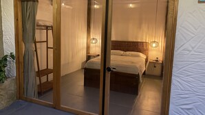 Ein Zimmer mit Bett, Nachttisch und Beistelltisch. Ein Fenster mit Vorhängen ist zu sehen, und in der Ecke steht eine Pflanze.