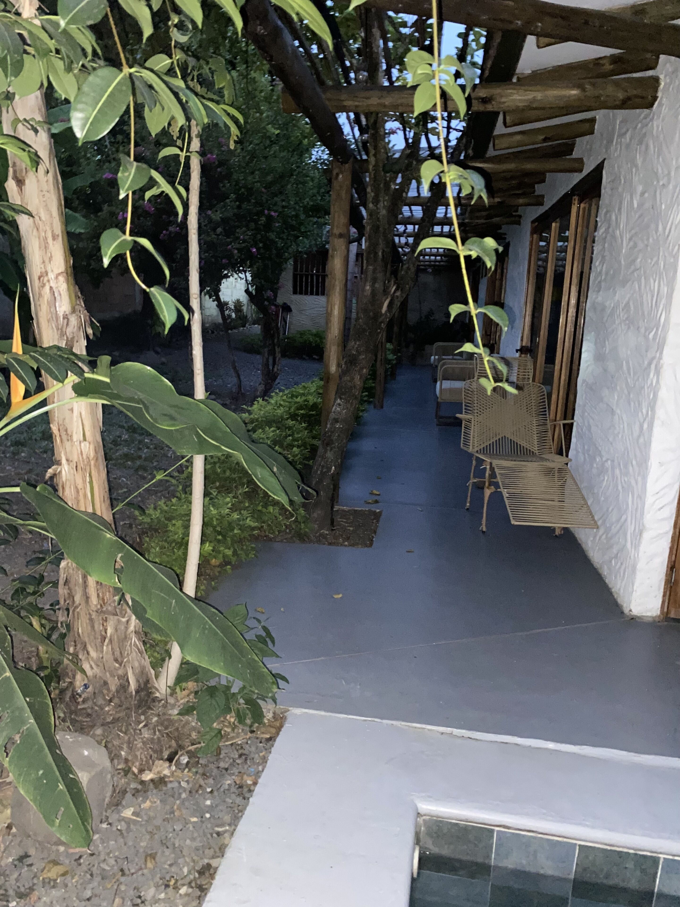 A narrow pathway leading to a building with a white exterior and a covered area with wicker furniture.