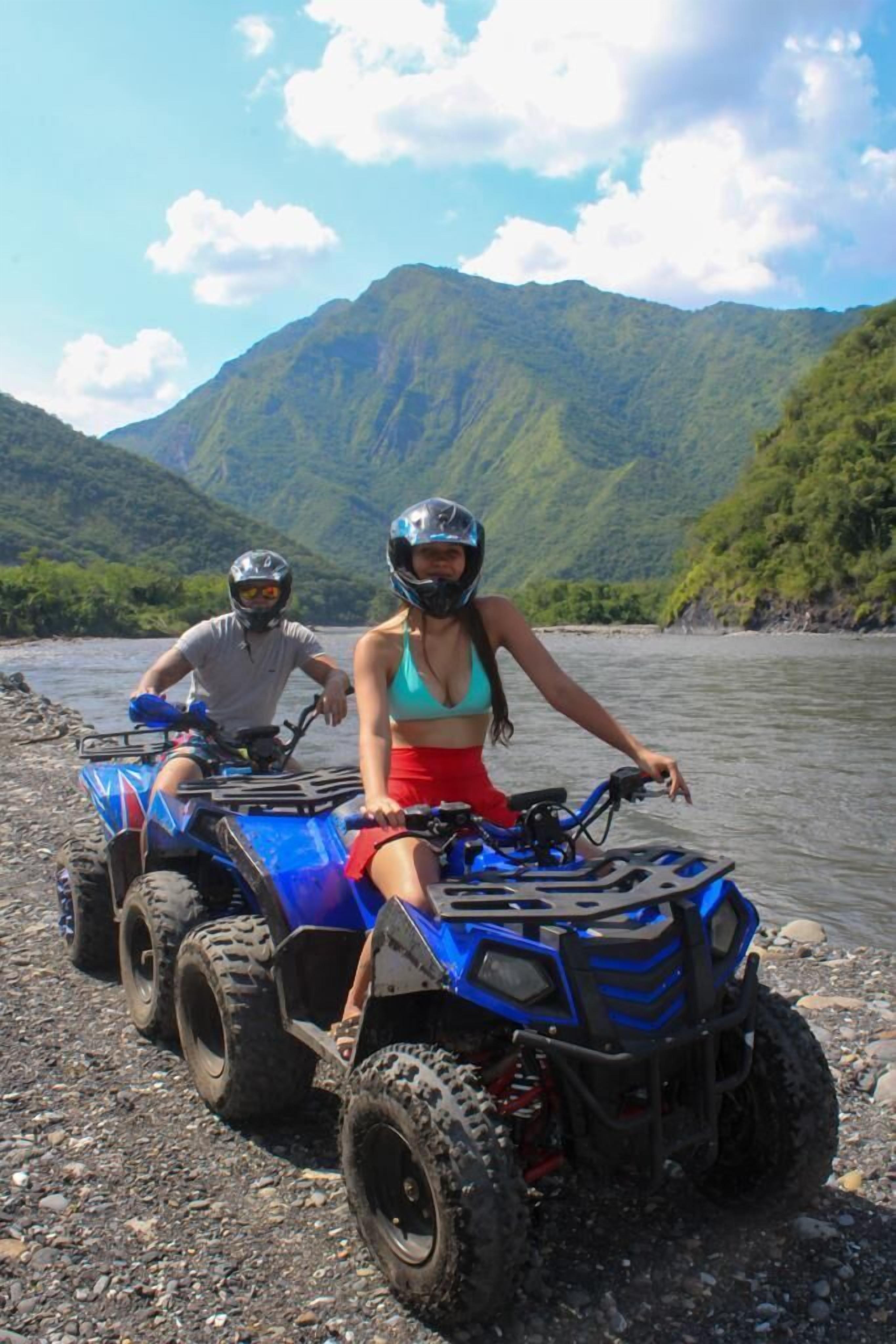 Dos personas en un cuatrimoto azul sobre la orilla rocosa de un río, con montañas al fondo.