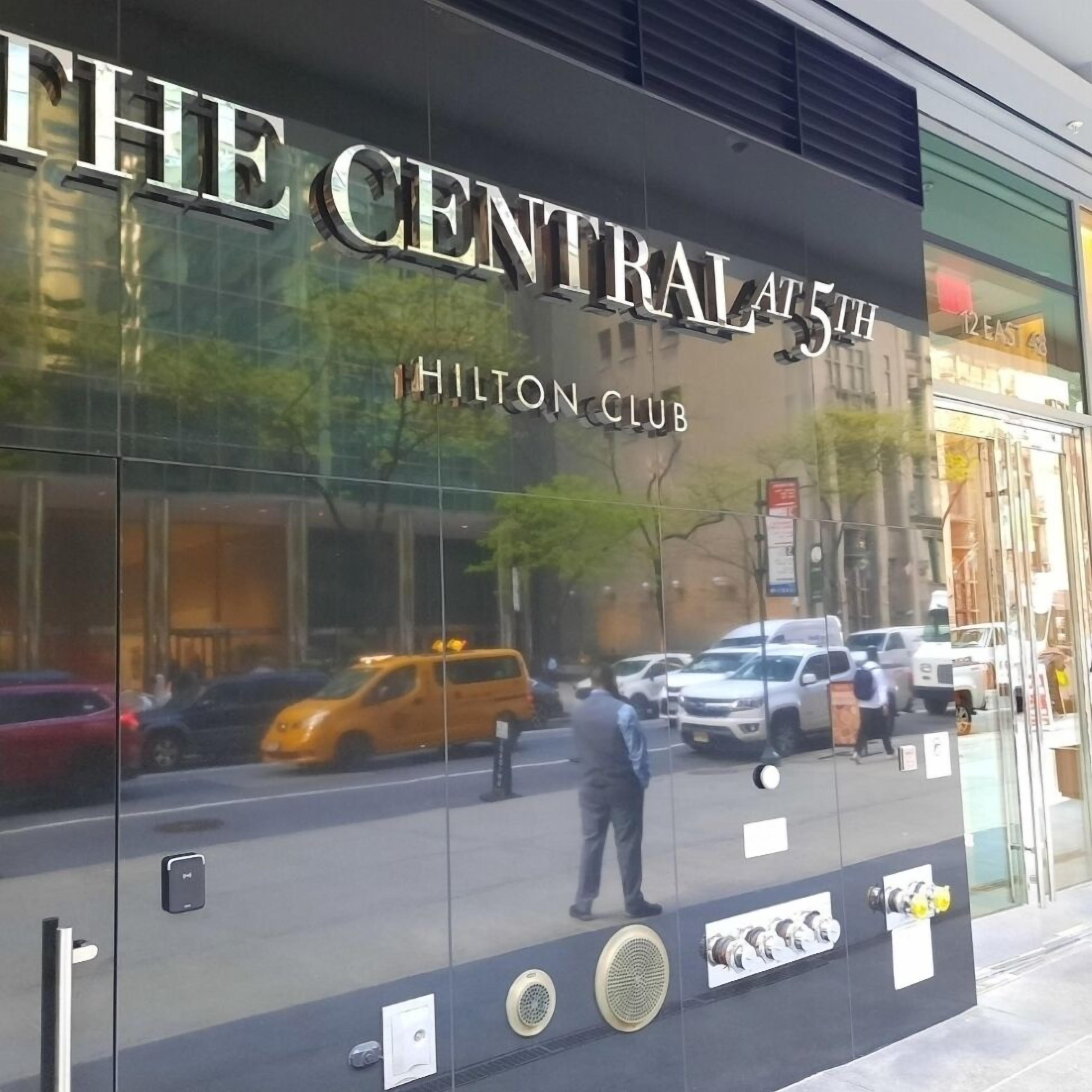 A glass facade of The Central at 5th Hilton Club with reflections of vehicles and a person on the ground.