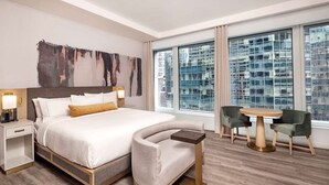 Una habitación de hotel moderna con una cama grande, un sillón curvo, una mesita pequeña y vistas de la ciudad a través de grandes ventanales.