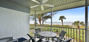 A covered balcony with a table and chairs, overlooking a beach with palm trees and the ocean.