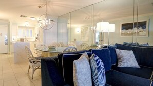 A modern living room with a dark blue sofa, a dining area with a glass table, and a large mirror reflecting the room.