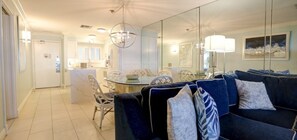 A modern living room with a dark blue sofa, a dining area with a glass table, and a large mirror reflecting the room.