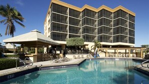 Un hotel con una piscina grande, una zona de estar techada y un edificio moderno con ventanales amplios.