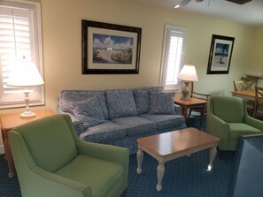 Uma sala de estar com um sofá estampado, uma poltrona verde, uma mesa de centro de madeira e quadros nas paredes.