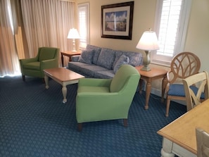 Quarto de hotel com carpete azul, poltrona verde, sofá, mesa de madeira e luminária.