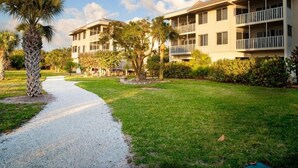Un chemin mène à un immeuble à plusieurs étages doté de balcons, entouré de pelouses bien entretenues et de palmiers.