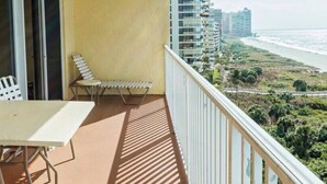 A balcony with a table and two chairs, overlooking a beach and high-rise buildings.