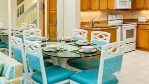 Un comedor con una mesa redonda de vidrio, sillas blancas con cojines azules y una cocina al fondo con armarios de madera y electrodomésticos blancos.