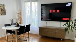 A modern living room with a flat-screen TV displaying Netflix, a dining area with a table and chairs, and a colorful rug on the wooden floor.