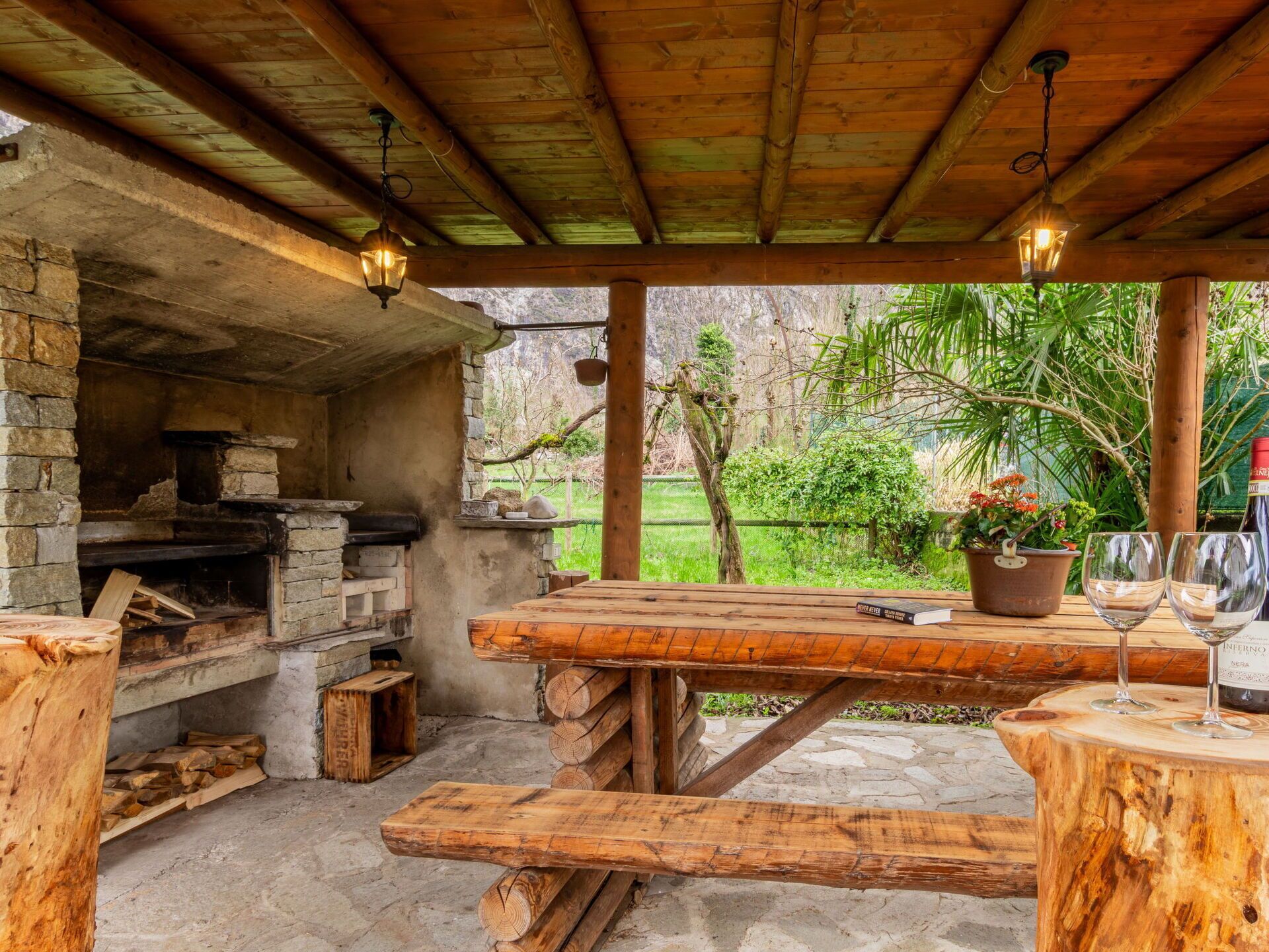 Une structure extérieure en bois dotée d’une cheminée en pierre, d’une table en bois avec des verres à vin et d’une plante en pot.