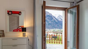 Ein Schlafzimmer mit einem Bett, einer Kommode, einer Lampe, einem Spiegel und Blick auf eine Berglandschaft und eine Stadt durch eine offene Tür.