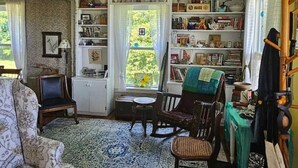 A cozy room with a wooden floor, a floral-patterned sofa, a rocking chair, a small table, and a bookshelf filled with books and decorative items. - Perry Shore Road Cottage (Perry)