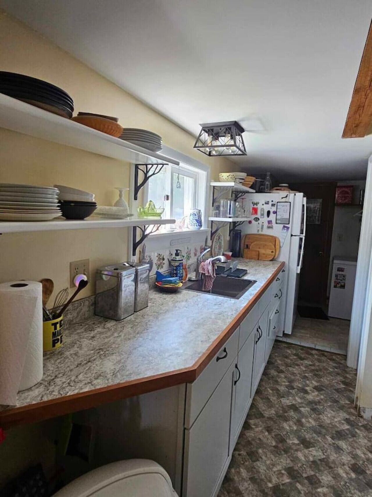 Une cuisine compacte avec des meubles blancs, un plan de travail en marbre et un réfrigérateur. Des étagères au-dessus du plan de travail servent à ranger la vaisselle, et un luminaire est suspendu au plafond.
