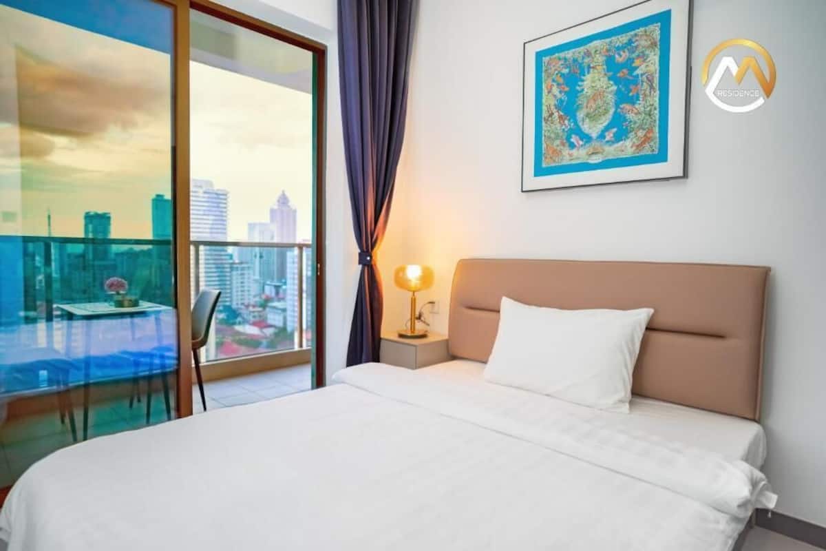 Habitación de hotel con cama, mesita de noche, lámpara, ventana con vista a la ciudad y un cuadro en la pared.