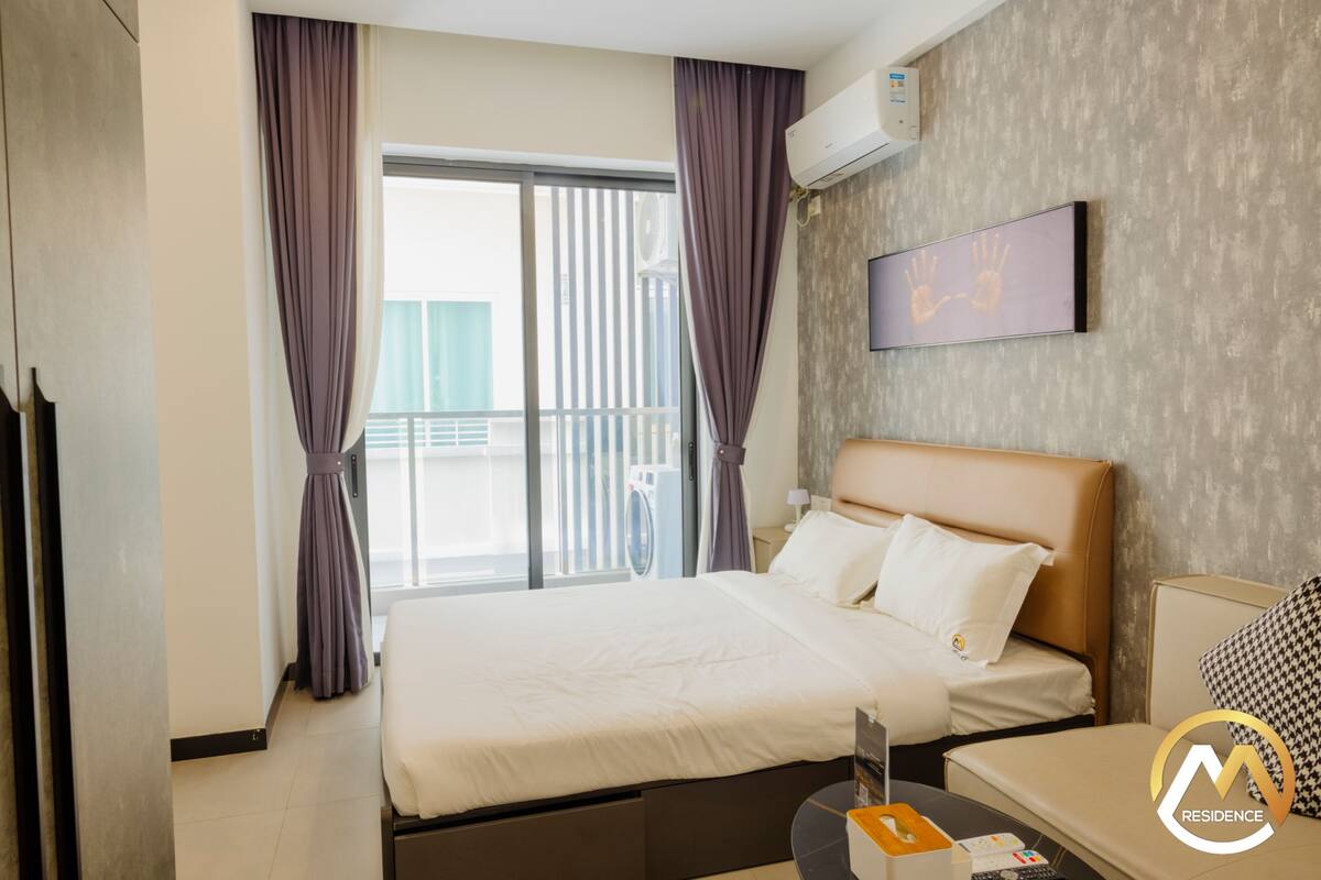 Habitación de hotel con cama, escritorio, silla, ventana con cortinas, equipo de aire acondicionado y una obra de arte colgada en la pared.