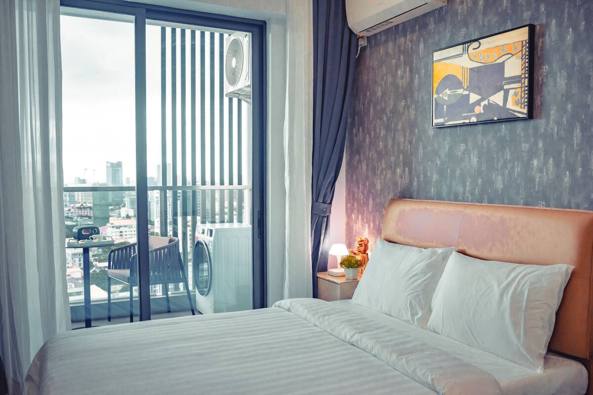 Habitación de hotel con una cama grande, una mesita de noche con lámpara y una planta pequeña, una ventana con cortinas y vistas a la ciudad.