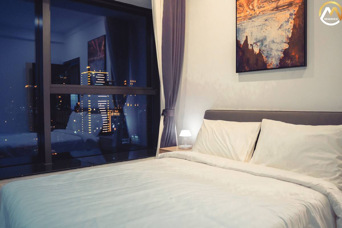 Una habitación de hotel con un ventanal grande, una cama, una mesita de noche, una lámpara y un cuadro en la pared.