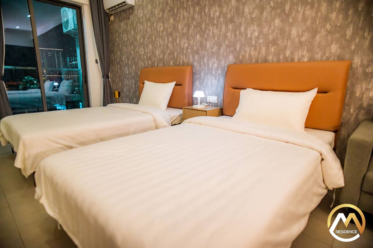 Habitación de hotel con dos camas, cada una con cabecero naranja y ropa de cama blanca. Hay una mesita de noche con lámpara y teléfono entre las camas.