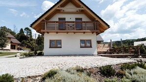 Une maison à deux étages dotée d’un balcon en bois, d’une allée pavée et d’un jardin planté de fleurs sauvages.