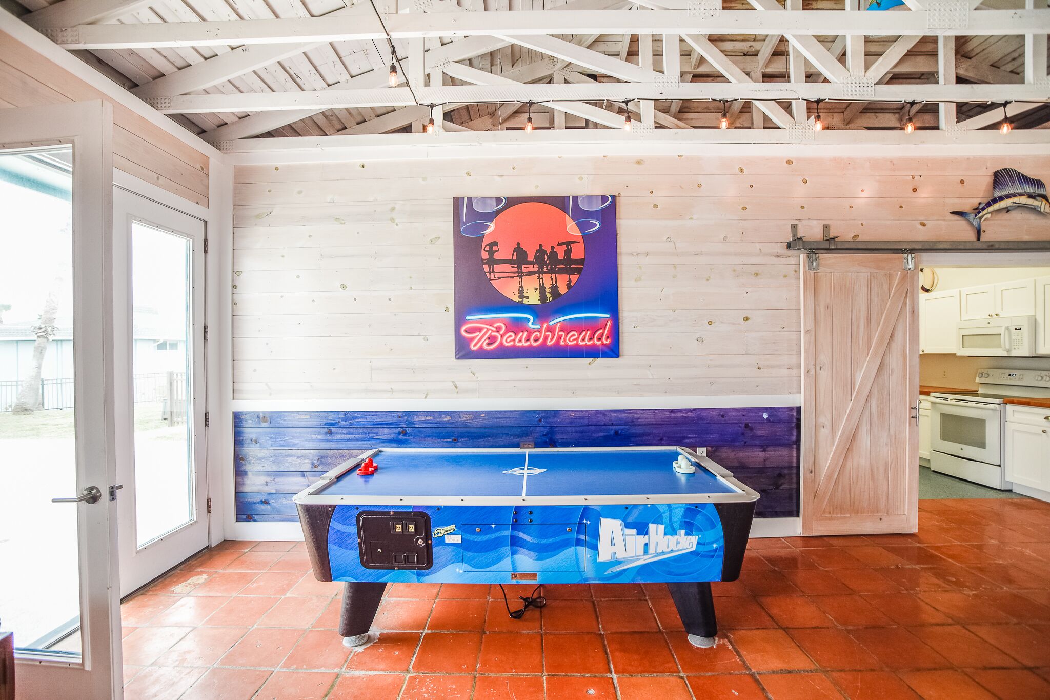 A pool table with a Beachhead logo, a kitchen area with white appliances, and a wooden ceiling with exposed beams.