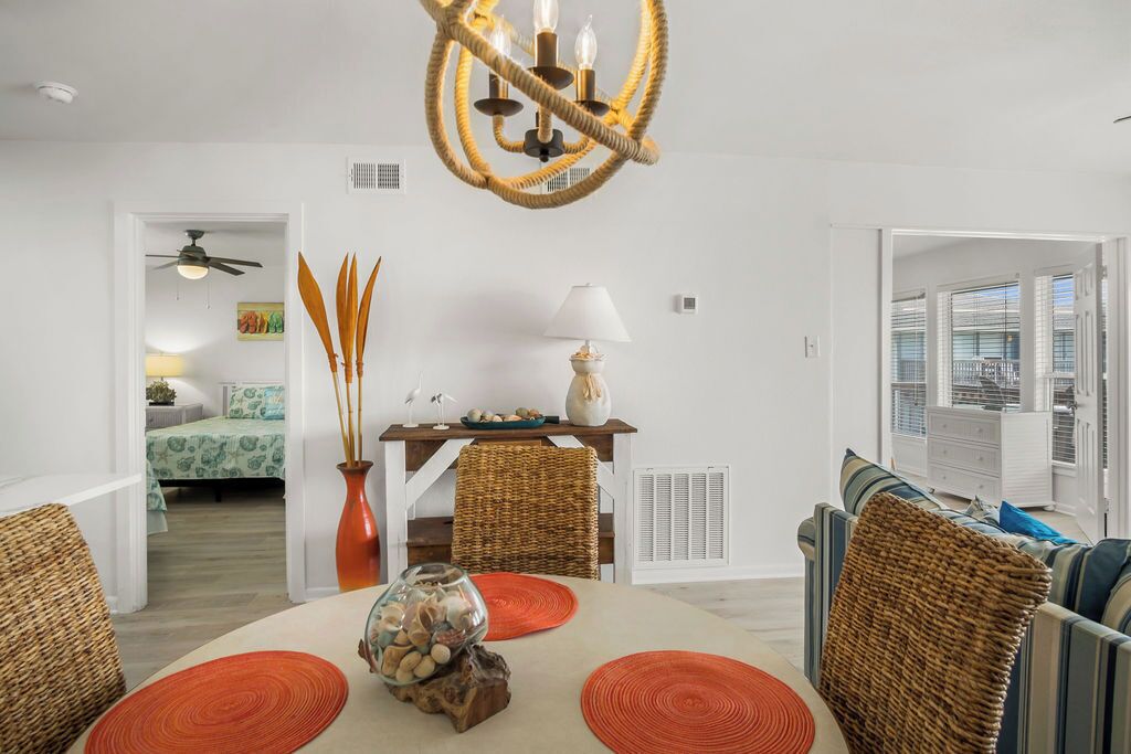 A dining area with a round table, orange placemats, wicker chairs, a chandelier, and a view of a bedroom with a bed and a ceiling fan.