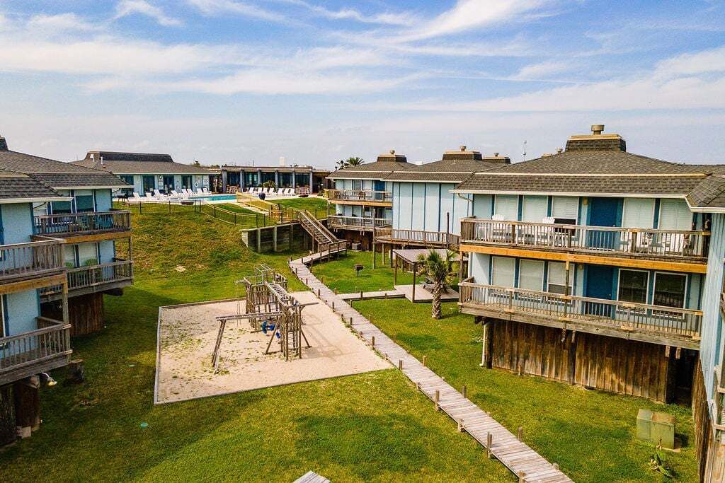 A beachfront hotel with multiple-story buildings, a sandy playground, and a wooden boardwalk.