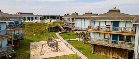 A beachfront hotel with multiple-story buildings, a sandy playground, and a wooden boardwalk.
