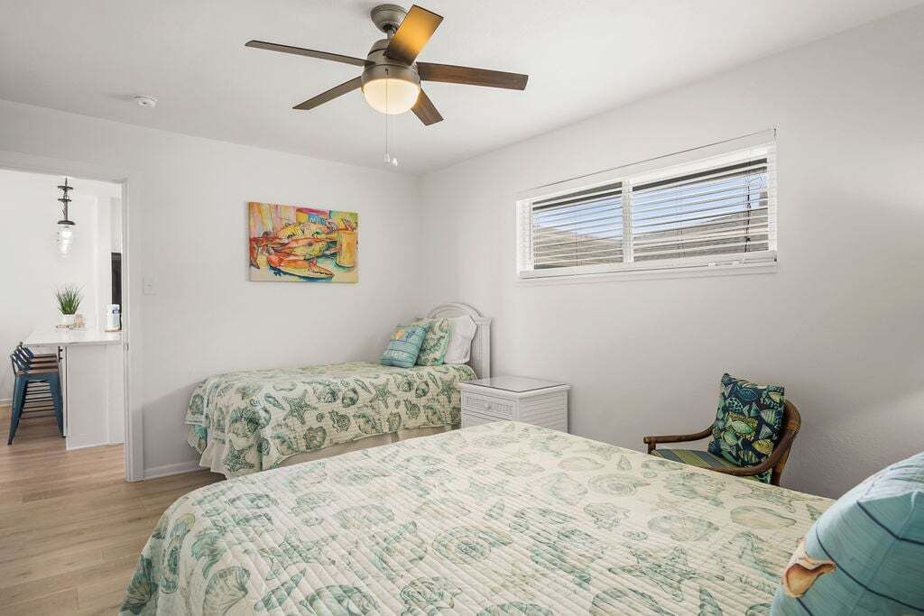 Ein Schlafzimmer mit zwei Betten, einem Deckenventilator, einem Fenster mit Jalousien, einem Schreibtisch und einem Stuhl.