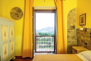Una habitación con cama, una mesita, un armario, un balcón con vistas a las montañas y una pared de piedra.