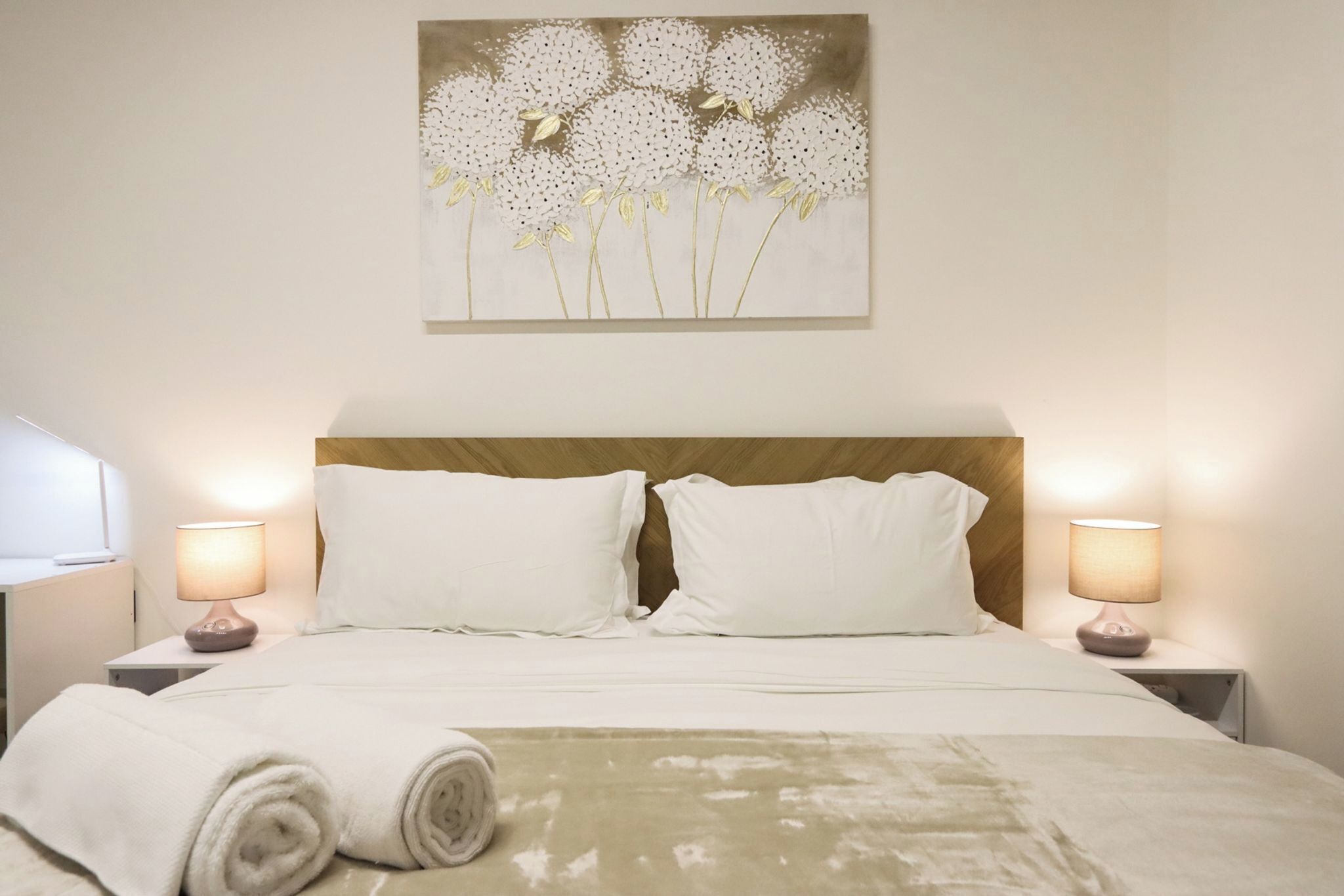 Un lit bien fait, avec du linge de lit blanc, deux lampes de chevet et une œuvre d’art représentant des fleurs accrochée au mur.